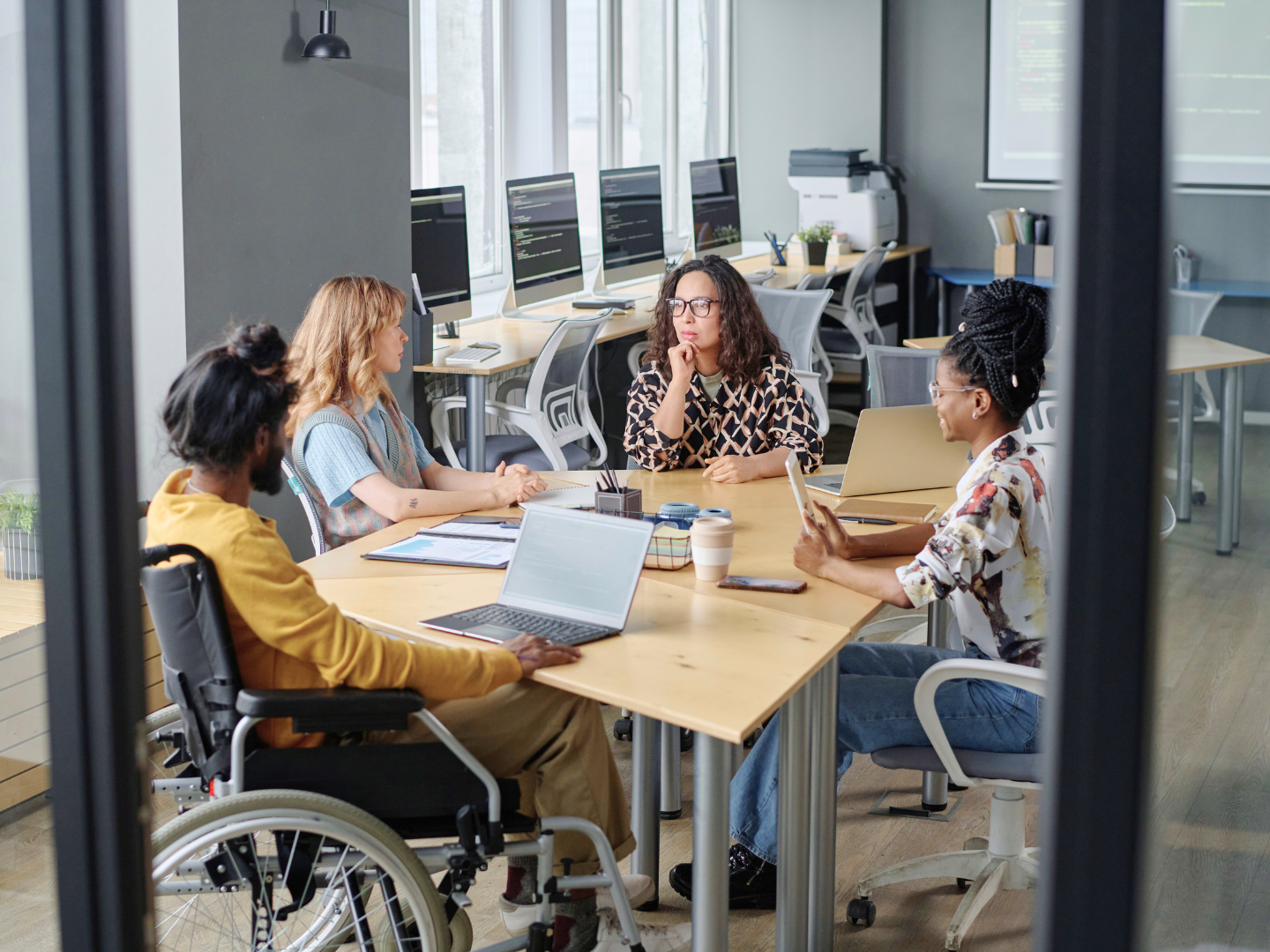 team of coworkers, a medium-skinned man in a wheelchair, a light-skinned young woman, a medium-skinned woman in glasses, and dark-skinned woman with braided hair sit around a conference table discussing how an S.B.A. loan can help their business grow