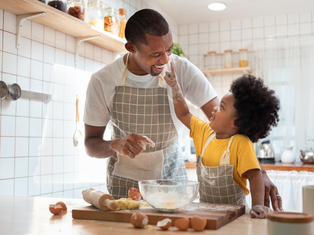 a father with dark skin and dark hair bakes in the kitchen of a home with financed maintained with personal checking accounts with a little boy with dark skin and dark curly hair 