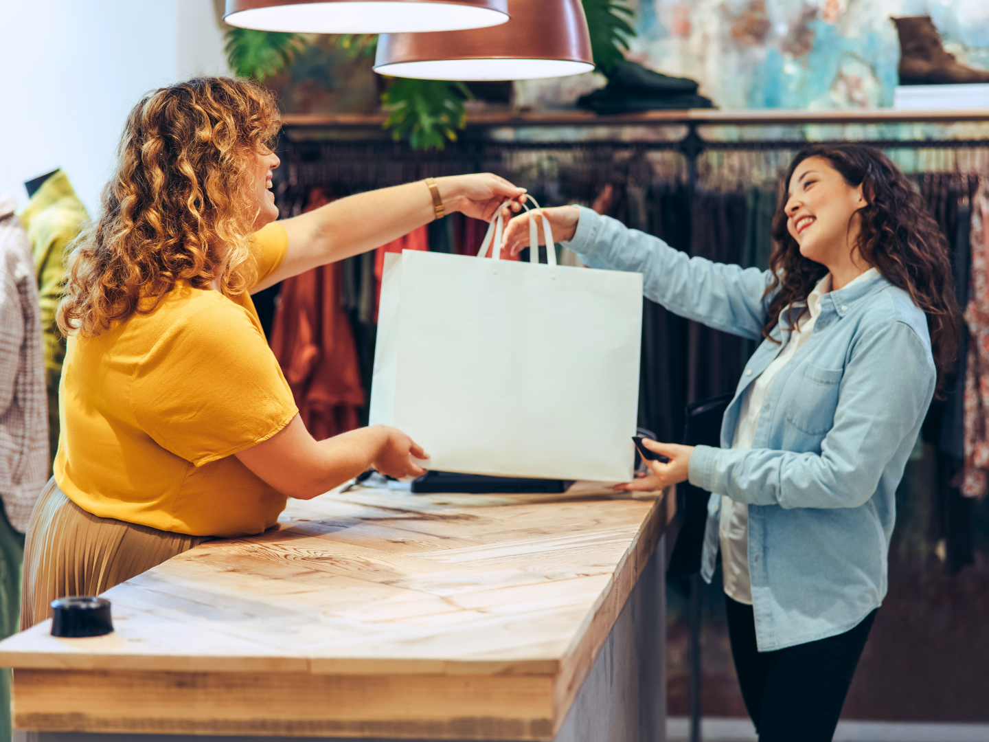 woman with dark curly hair and medium skin accepts a shopping bag from a boutique owners where she has purchased clothing using funds in her personal checking account