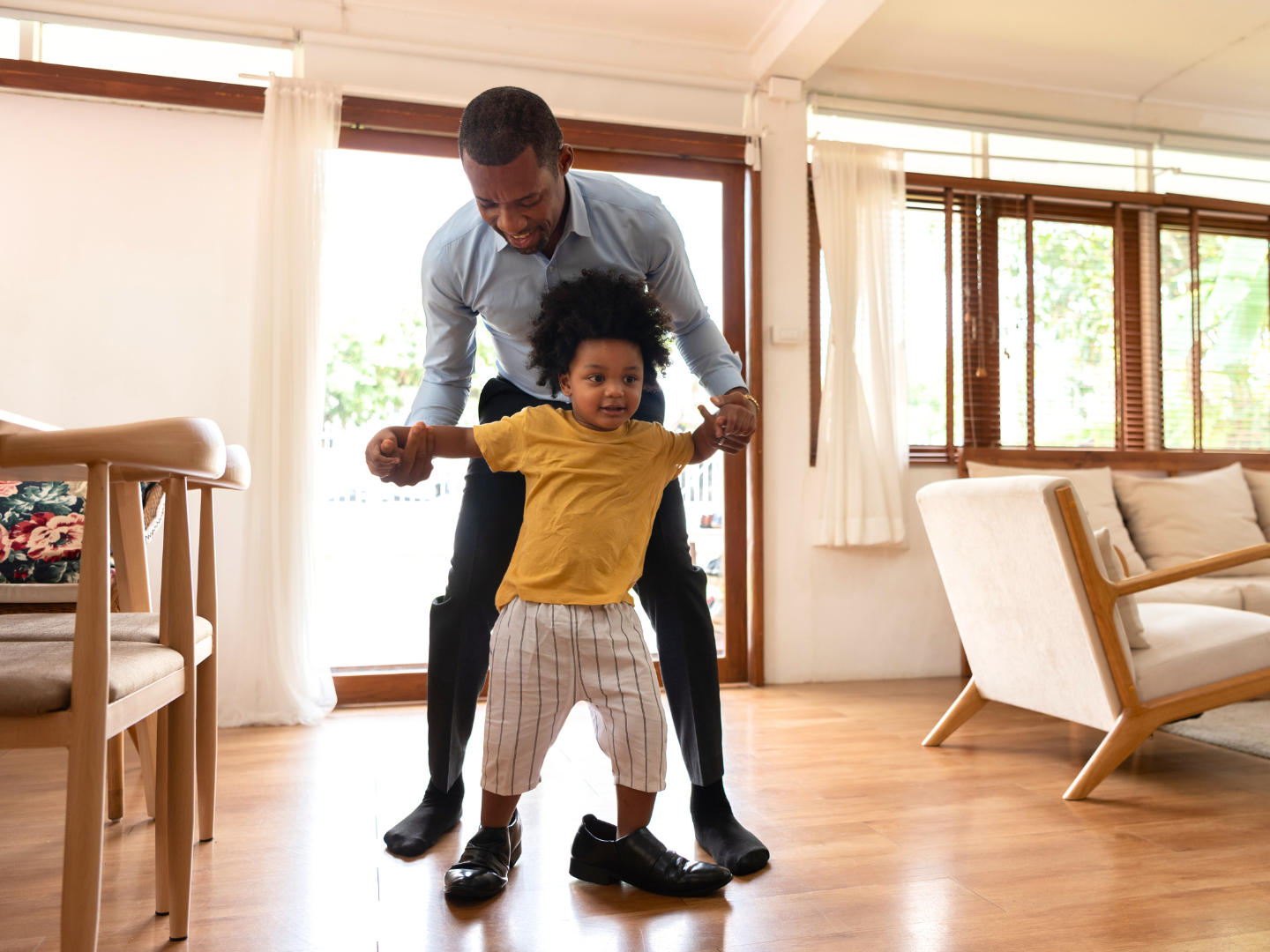 a single father with dark skin and dark hair in office attire holds the hands of a toddler boy playing in his fathers shoes after making plans to protect his savings in an unstable economy