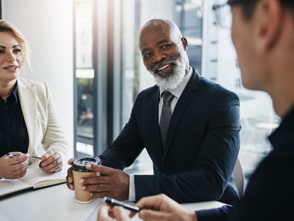 a male business owner with brown skin and a gray beard wearing a suit sits with his employees discussing the benefits of minority business financing